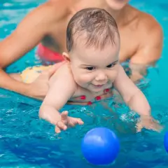 Glad bebis med ett leende i vattnet, en person håller i den. Två blå bollar flyter framför. Perfekt för babysim ålder.