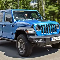 Niebieski Jeep Wrangler 4xe Rubicon z kierowcą za kierownicą jedzie po drodze wśród drzew.