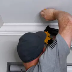 A man is performing crown molding installation, using a nail gun to secure the decorative trim to the ceiling.