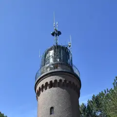 Ceglana łeba latarnia morska z metalową galeryjką i antenami na szczycie, otoczona sosnami, na tle błękitnego nieba.