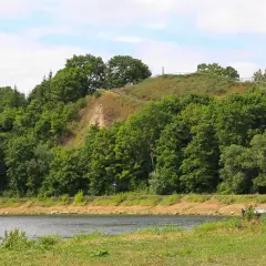 Zielona skarpa nad rzeką, na szczycie której widać ścieżki i ogrodzenie. To malownicza góra zamkowa w Drohiczynie.