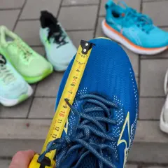 Miarka mierzy niebieskie buty do biegania. Czy buty do biegania powinny być większe? W tle inne kolorowe buty.