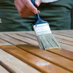 Application d'une lasure acrylique ou glycéro sur des lattes de bois. La brosse dépose le produit, transformant le bois clair en une teinte plus foncée et riche.