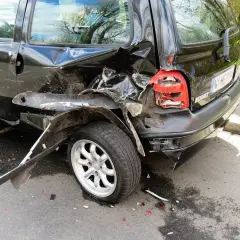 Czarny samochód po kolizji, z widocznymi uszkodzeniami tylnej części i koła.