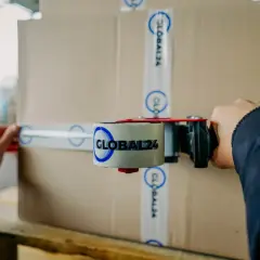Ręce zamykają karton taśmą z logo GLOBAL24, przygotowując paczkę do hermes przesyłki zagraniczne.
