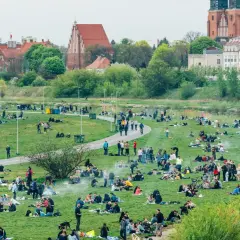 Ludzie odpoczywają na trawie w nadwarciańskiej części Poznania. W tle widać zabytkowe budynki i rzekę.