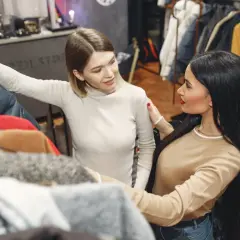 Dwie kobiety przeglądają ubrania w sklepie. Jedna z nich dotyka kurtki, druga wspiera ją ramieniem. W tle widać wieszaki z odzieżą, sugerujące skup ubrań używanych.