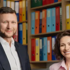 Uśmiechnięci mężczyzna w garniturze i kobieta w różowej koszuli na tle regałów z kolorowymi segregatorami.