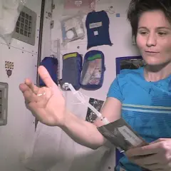 Astronautka pokazuje kroplę wody w dłoni, która unosi się w przestrzeni kosmicznej. Trzyma też opakowanie z napisem "Drinking Water".
