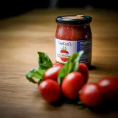 Barattolo di pomodoro siccagno pizzutello pelato in succo, con pomodorini freschi e basilico su un tavolo di legno.