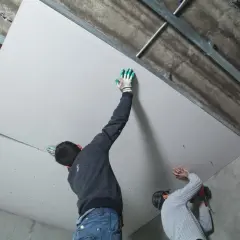 Deux ouvriers s'affairent à habiller un plafond abîmé avec des plaques de plâtre, fixées sur une structure métallique.