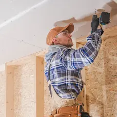 Operaio con trapano avvita pannelli per insonorizzazione stanza. Struttura in legno e cartongesso visibile.