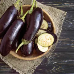 Einige ganze Auberginen liegen neben geschnittenen Scheiben in einer Holzschale. Man sieht das helle Fruchtfleisch der Aubergine innen.