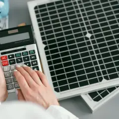 Ręce liczą koszty inwestycji w przemysłowe magazyny energii przy pomocy kalkulatora, obok panele słoneczne i skarbonka.