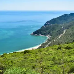 Costa verde en el sur de Lisboa, con mar turquesa, playa, barcos y montañas cubiertas de vegetación.