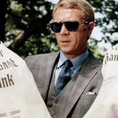 Steve McQueen w okularach przeciwsłonecznych, w eleganckim garniturze, trzyma worki z napisem "Mercantile Bank". Scena z filmu "Afera Thomasa Crowna".