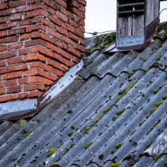 Stary dach sąsiada pokryty eternitem, który może zawierać azbest, porośnięty mchem. Obok ceglany komin.