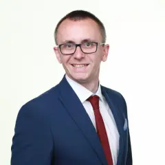 Mężczyzna w okularach, garniturze i krawacie, uśmiecha się na białym tle.