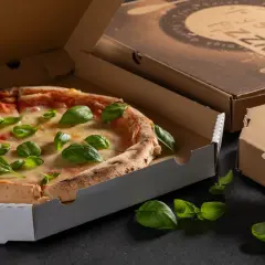 Pyszna pizza w otwartym opakowaniu na pizze, ozdobiona świeżą bazylią. Obok zamknięte kartony.