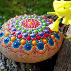Bunte bemalte Steine mit Mandala-Muster, die auf einem Baumstamm liegen, umgeben von grünem Gras und gelben Blüten.