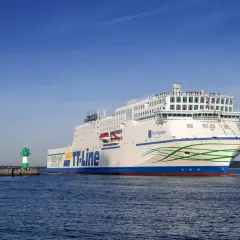 Duży biały prom TT-Line "Nils Holgersson" płynie po wodzie obok zielono-białej latarni morskiej.
