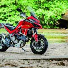 Czerwony Ducati Multistrada 1200S zaparkowany na piaszczystym brzegu rzeki, otoczony bujną zielenią.