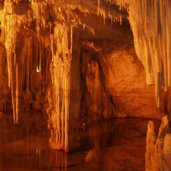 Interior de la cueva Rei Cintolo, con estalactitas y estalagmitas iluminadas en tonos cálidos, reflejándose en el agua subterránea.