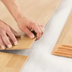 Hands installing laminate flooring over a subfloor. A stack of laminate planks is visible on the right. This shows how you can put laminate flooring over vinyl.