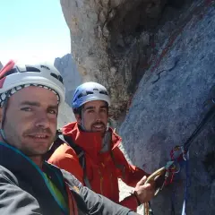 Dos escaladores sonríen en la cara del Naranjo de Bulnes, equipados con cascos y arneses, listos para la aventura.