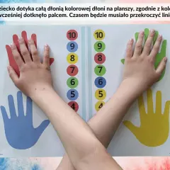 Dziecko wykonuje ćwiczenia na przekraczanie linii środka, dotykając dłonią kolorowych dłoni na planszy.