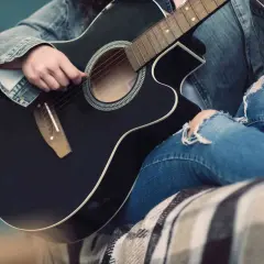 Osoba w dżinsowej kurtce i podartych jeansach gra na czarnej gitarze. Słychać delikatne dźwięki, jakby tworzyła własną melodię.
