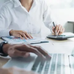 Dwie osoby pracujące przy biurku z dokumentami, kalkulatorem i laptopem.