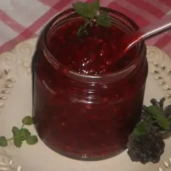 Słoik domowej frużeliny malinowej, z łyżeczką zanurzoną w gęstym, czerwonym musie. Udekorowana listkami mięty i szyszkami.