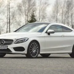 Biały Mercedes C klasa coupe z czarnym dachem, zaparkowany na brukowanej nawierzchni, z drzewami w tle.