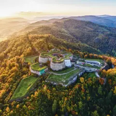 Twierdza na wzgórzu, otoczona jesiennymi lasami. W oddali widać pasmo górskie.