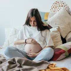 Kobieta w ciąży, otoczona poduszkami, czule dotyka brzucha. Czas oczekiwania na dziecko, być może zmagająca się z wielowodziem, ale pełna spokoju.