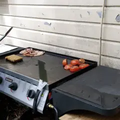 Une main utilise des pinces pour retourner des saucisses sur une plancha à gaz. Des tranches de bacon et des tomates grillées sont également présentes.