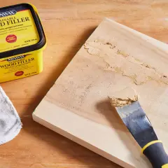 Un pot de mastic à bois Minwax est à côté d'une planche de bois où un couteau à mastic applique le produit pour reboucher bois.