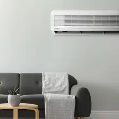 Unidad de aire acondicionado montada en la pared, cumpliendo la normativa aire acondicionado. Sofá gris con mantas y mesa auxiliar con planta.