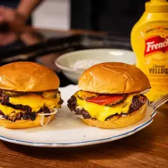 Två saftiga smashed burgers med smält ost, pickles och lök. Bredvid står flaskor med senap och ketchup.