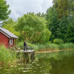 Dwaj wędkarze na pomoście przy czerwonej chacie, otoczeni zielenią. Idealne miejsce na Kaszubach dla miłośników spokoju.