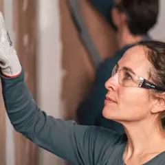 Donna con occhiali protettivi usa una levigatrice per imparare come levigare un muro.