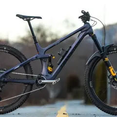 A dark purple full-suspension mountain bike with yellow accents sits on a road. Its components suggest a focus on performance, hinting at a competitive mountain bike weight for agile trail riding.