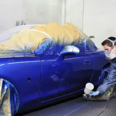 Un carrozziere con maschera spruzza vernice blu su un'auto coperta. Consulta il listino prezzi verniciatura auto per i costi.