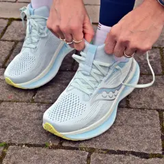 Dłonie zawiązują sznurówki jasnoniebieskich butów do biegania Saucony Ride 18 na chodniku z kostki brukowej.