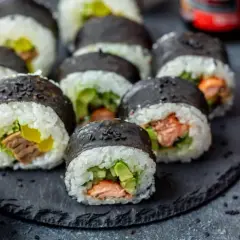 Gotowe do jedzenia rolki sushi z łososiem i awokado, obok słoiczek marynowanego imbiru i miseczka czarnego sezamu – idealne składniki do sushi.