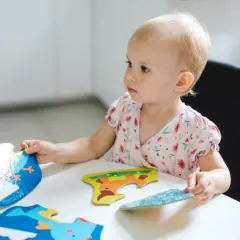 Mała dziewczynka z blond włosami skupiona na układaniu kolorowych puzzli z zwierzątkami.