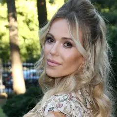Doda IQ, blondynka w kwiecistej sukience, z burzą loków, uśmiecha się do obiektywu na tle zieleni.