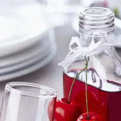 Elegancka butelka z wiśniowym alkoholem, ozdobiona wstążką i świeżymi wiśniami, obok kieliszka i stosu talerzy.