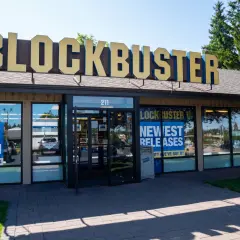 L'insegna dorata del negozio Blockbuster domina l'edificio. Vetrine mostrano "Best Selection" e "Newest Releases", un richiamo nostalgico ai tempi d'oro del blockbuster film.